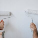 Dwie osoby malują białą ścianę przy użyciu wałków malarskich.