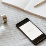 Smartfon z napisem "have less, dom more" na ekranie, notatnik, długopis i butelka wody na prześcieradle.